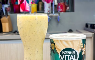 Fotografia em tons de verde e azul em uma bancada de cozinha com um copo alto de vidro e a vitamina de maracujá, banana e coco dentro dele.