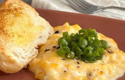 a imagem mostra uma porção de ovos mexidos em tons de amarelo, cobertos por cebolinhas picadas e uma torrada ao lado. Ele está em um prato oval de cor marrom avermelhada, com um guardanapo de pano bege e um garfo atrás.