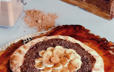 Fotografia em tons de marrom, em uma bancada de madeira de cor branca. Ao centro, uma toalha colorida azul com uma tábua de madeira redonda em cima. Em cima de tábua, está a pizza e ao fundo, há um pote com canela em pó. Fotografia em tons de marrom, em uma bancada de madeira de cor branca. Ao centro, uma toalha colorida azul com uma tábua de madeira redonda em cima. Em cima de tábua, está a pizza e ao fundo, há um pote com canela em pó.