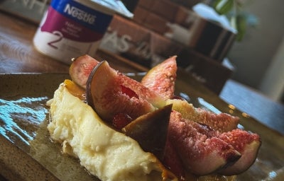 Fotografia de uma receita de iogurte assado com pedaços de figos por cima. A sobremesa está em um apoio retangular, e ao fundo tem uma embalagem do NESTLÉ Iogurte Natural Zero Lactose.