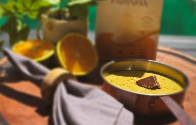 Fotografia de uma receita em tons claros sob o sol de uma mini panelinha com mingau de laranja e um quadradinho de chocolate decorando, por cima. Ao redor tem um panino cinza, duas metades de uma laranja e uma embalagem de NESTLÉ Farinha de Aveia.