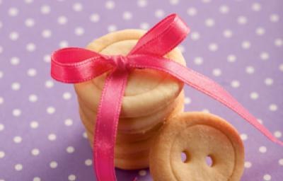 fotografia em tons de roxo, bege e rosa de uma bancada roxa com bolinhas brancas, contém biscoitinhos em formatos de botão com um laço rosa por cima. fotografia em tons de roxo, bege e rosa de uma bancada roxa com bolinhas brancas, contém biscoitinhos em formatos de botão com um laço rosa por cima.