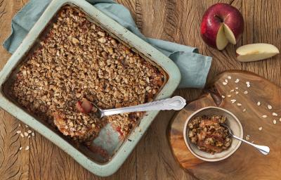 Fotografia em tons de azul em uma mesa de madeira escura com uma travessa retangular azul e o crumble de maçã e aveia dentro. Ao lado, uma tábua de madeira e um potinho com um pedaço do crumble. Ao fundo, uma maçã cortada ao meio. Fotografia em tons de azul em uma mesa de madeira escura com uma travessa retangular azul e o crumble de maçã e aveia dentro. Ao lado, uma tábua de madeira e um potinho com um pedaço do crumble. Ao fundo, uma maçã cortada ao meio.