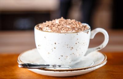 fotografia em tons de branco e marrom de uma bancada marrom vista de frente. Ao centro contém um pratinho redondo branco com uma xicara branca por cima, dentro da xicara contém a bebida e por cima raspas de chocolate, ao lado uma colher para mexer. fotografia em tons de branco e marrom de uma bancada marrom vista de frente. Ao centro contém um pratinho redondo branco com uma xicara branca por cima, dentro da xicara contém a bebida e por cima raspas de chocolate, ao lado uma colher para mexer.