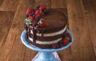 Imagem da receita de Naked Cake de Cacau, decorado em camadas marrons e brancas, com chocolate escorrendo, decorado com frutas vermelhas. O bolo está sobre um prato azul em uma bancada de madeira. Imagem da receita de Naked Cake de Cacau, decorado em camadas marrons e brancas, com chocolate escorrendo, decorado com frutas vermelhas. O bolo está sobre um prato azul em uma bancada de madeira.