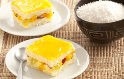 Fotografia em tons de amarelo em uma bancada de madeira com uma toalha de palha, dois pratos brancos redondos com um pedaço de pavê de tapioca em cada um. Ao lado, um potinho preto com coco ralado. Fotografia em tons de amarelo em uma bancada de madeira com uma toalha de palha, dois pratos brancos redondos com um pedaço de pavê de tapioca em cada um. Ao lado, um potinho preto com coco ralado.