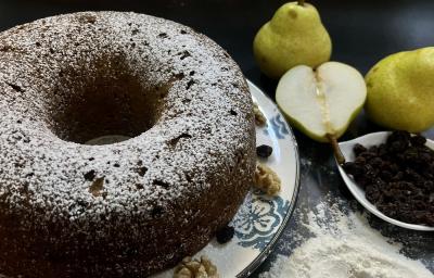 Foto da receita de bolo de pera com aveia em tons escuros servida em um prato de porcelana decorado sobre uma mesa com toalha adornada em azul e branco. Ao fundo, duas eras inteiras e uma metade como decoração, além de um recipiente com uvas passas.