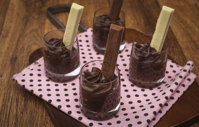 Fotografia em tons de rosa em uma bancada de madeira, uma tábua de madeira com um pano rosa e bolinhas pretas, quatro copinhos de vidro com brigadeiro de colher de chocolate dentro de cada um com um KitKat ao Leite e KitKat Branco dentro dos copinhos. Fotografia em tons de rosa em uma bancada de madeira, uma tábua de madeira com um pano rosa e bolinhas pretas, quatro copinhos de vidro com brigadeiro de colher de chocolate dentro de cada um com um KitKat ao Leite e KitKat Branco dentro dos copinhos.