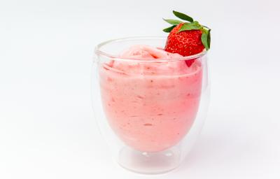 Fotografia de um creme de morango com erva cidreira em um recipiente de vidro de decoração sobre uma mesa branca. Em cima do creme, há um morango inteiro decorando.
