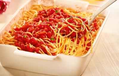 Fotografia em tons de marrom, amarelo e vermelho de uma bancada de madeira clara, sobre ela um recipiente quadrado branco com macarrão, molho sugo e uma colher de servir com uma porção do macarrão. Ao fundo um guardanapo vermelho com listras brancas. Fotografia em tons de marrom, amarelo e vermelho de uma bancada de madeira clara, sobre ela um recipiente quadrado branco com macarrão, molho sugo e uma colher de servir com uma porção do macarrão. Ao fundo um guardanapo vermelho com listras brancas.