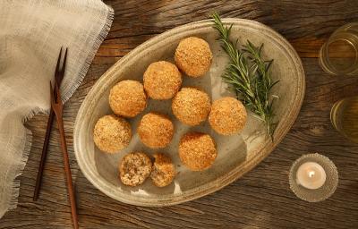 Foto em tons de marrom da receita de arancini de aveia servido em diversas porções sobre uma tábua circular de cerâmica com um ramo de manjericão, em cima de uma mesa de madeira e ao lado dois garfos de madeira sobre um pano bege de linho
