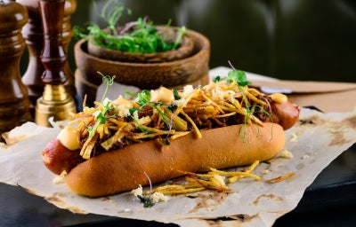 Imagem mostra um cachorr quente com pão, salsicha, batata-palha, molhos e ervas verdes