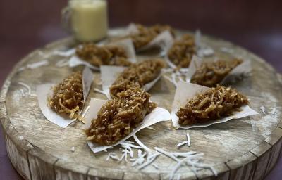 Foto da receita de cocada com doce de leite em tons de marrom servidas em oito porções em lenços de papel sobre uma tábua de maderia redonda e grossa. Ao fundo, há um recipiente de vidro com leite MOÇA para decorar Foto da receita de cocada com doce de leite em tons de marrom servidas em oito porções em lenços de papel sobre uma tábua de maderia redonda e grossa. Ao fundo, há um recipiente de vidro com leite MOÇA para decorar