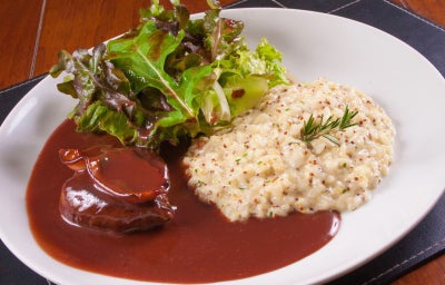 Fotografia em tons claros com uma receita de arroz a piamontese ao centro acompanhada de carne e folhas.