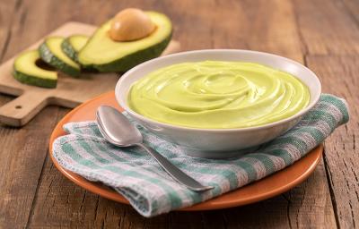 Foto em tons de verde da receita de creme rústico de abacate servida em um bowl de porcelana branco com uma colher prateada ao lado sobre um pano verde e branco quadriculado, tudo sobre um prato de porcelana laranja. Ao fundo, tábua com fatias de abacate Foto em tons de verde da receita de creme rústico de abacate servida em um bowl de porcelana branco com uma colher prateada ao lado sobre um pano verde e branco quadriculado, tudo sobre um prato de porcelana laranja. Ao fundo, tábua com fatias de abacate