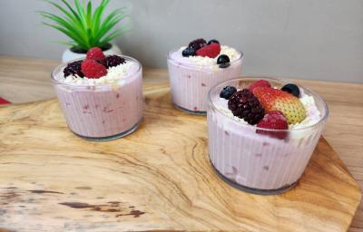 Fotografia em tons de rosa em uma bancada de madeira clara com três potes de vidro e mousse dentro de cada um deles. Frutas vermelhas decorando.