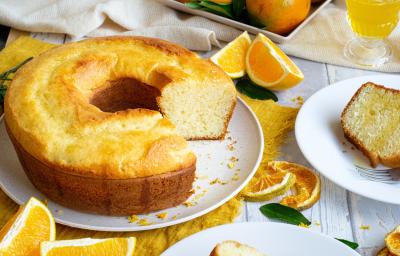 Fotografia de um bolo de laranja feito em forma de furo central sobre um prato raso de cerâmica branco. Ao lado tem um prato de sobremesa com uma fatia do bolo e um garfo, e ao redor tem pedaços e fatias de laranja, sobre uma mesa de madeira branca. Fotografia de um bolo de laranja feito em forma de furo central sobre um prato raso de cerâmica branco. Ao lado tem um prato de sobremesa com uma fatia do bolo e um garfo, e ao redor tem pedaços e fatias de laranja, sobre uma mesa de madeira branca.