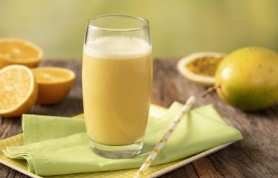 Fotografia em tons de amarelo, verde e marrom de uma bancada de madeira com uma bandeja amarela e um paninho verde com um canudo sobre ele um copo de vidro com a vitamina de laranja. Ao fundo três metades de laranjas, um maracujá inteiro e uma metade. Fotografia em tons de amarelo, verde e marrom de uma bancada de madeira com uma bandeja amarela e um paninho verde com um canudo sobre ele um copo de vidro com a vitamina de laranja. Ao fundo três metades de laranjas, um maracujá inteiro e uma metade.