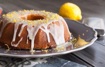 Fotografia de um bolo de limão em uma elegante travessa, coberto com glacê branco que escorre pelas laterais. Raspas de limão decoram o topo, enquanto um limão inteiro e um ralador estão ao fundo.