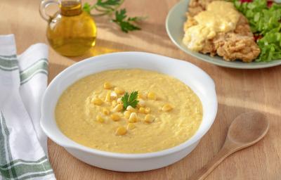 Foto da Receita de Creme de Milho Sem Lactose. Observa-se um recipiente branco com o creme de milho dentro e, atrás, um prato de filé à milanesa servido com o creme