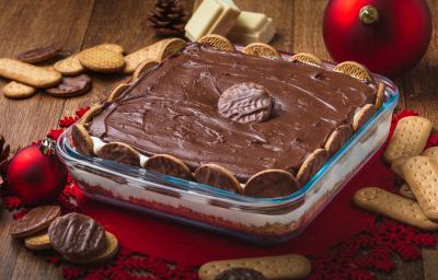 Foto da receita de Torta Holandesa na Travessa, em uma bancada de madeira decorada com temas de natal, alguns biscoitos espalhados, pedaços de chocolate branco. A receita tem um creme marrom sobre um creme claro e está em um refratário de vidro