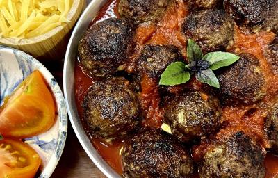 Foto em tons de vermelho escuro da receita de almôndegas recheadas servida em um prato grande e fundo de porcelana branco. Ao lado dois pratos menores, um com tomates e outro com queijo ralado. Foto em tons de vermelho escuro da receita de almôndegas recheadas servida em um prato grande e fundo de porcelana branco. Ao lado dois pratos menores, um com tomates e outro com queijo ralado.