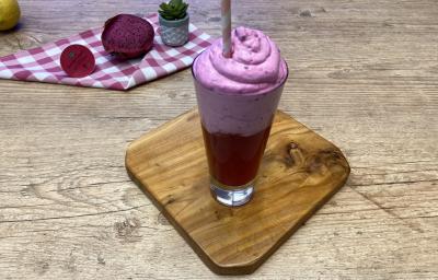 Fotografia em tons de rosa em uma bancada de madeira clara com uma tábua de madeira ao centro e um copo de vidro alto com uma bebida rosa e uma espuma rosa. Ao fundo, um pano xadrez rosa com uma pitaya aberta na metade e um limão-siciliano. Fotografia em tons de rosa em uma bancada de madeira clara com uma tábua de madeira ao centro e um copo de vidro alto com uma bebida rosa e uma espuma rosa. Ao fundo, um pano xadrez rosa com uma pitaya aberta na metade e um limão-siciliano.