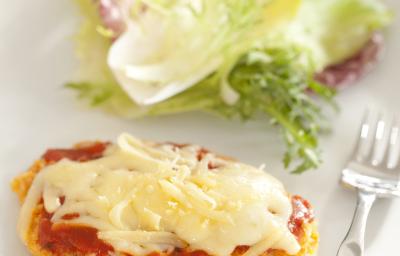 Fotografia em tons de marrom em uma bancada de madeira com uma toalha cinza, um sousplat marrom, um prato branco redondo com o filé à parmegiana acompanhado de salada. Ao fundo, um potinho de azeite. Fotografia em tons de marrom em uma bancada de madeira com uma toalha cinza, um sousplat marrom, um prato branco redondo com o filé à parmegiana acompanhado de salada. Ao fundo, um potinho de azeite.