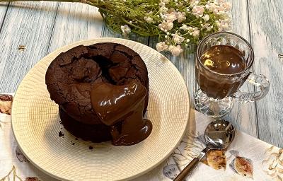 Foto em tons de bege e marrom da receita de bolo fudge servida sobre um prato bege e uma tolha de mesa decorada. Ao lado, há uma colher prateada e uma caneca de vidro com chocolate derretido. Ao fundo, um buqué de flores cor de rosa Foto em tons de bege e marrom da receita de bolo fudge servida sobre um prato bege e uma tolha de mesa decorada. Ao lado, há uma colher prateada e uma caneca de vidro com chocolate derretido. Ao fundo, um buqué de flores cor de rosa