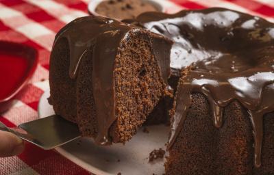 Foto bem aproximada de um bolo de chocolate com uma fatia sendo retirada. O bolo está num prato branco e coberto com Brigadeiro e o prato está sobre uma toalha xadrez vermelho e branco. Foto bem aproximada de um bolo de chocolate com uma fatia sendo retirada. O bolo está num prato branco e coberto com Brigadeiro e o prato está sobre uma toalha xadrez vermelho e branco.
