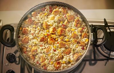 Foto de cima de uma panela com a receita de arroz caipira, em tons branco e amarelo, com pedaços de frango e milho. A panela está sobre um fogão e dela sai uma fumaça.
