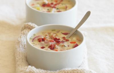 Fotografia de dois recipientes de sopa fundos e na cor branca, com um caldo de batata e salsão com cenoura, bacon, cebola roxa e massa. O recipiente da frente está apoiado em uma toalha de lá na cor bege, que está sobre uma mesa na cor bege claro.