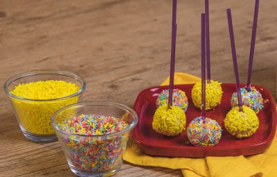 Fotografia em tons de amarelo e vermelho em uma bancada de madeira clara, um pano amarelo e um prato vermelho com alguns pirulitos de ninho com granulado colorido em cima dele. Ao lado, potinhos com os granulados coloridos e a lata de Ninho em Pó.