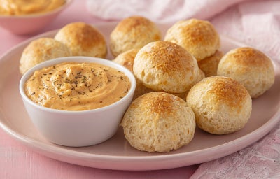 Imagem mostra uma porção de pães de queijo dourados com um recipiente transparente contendo um creme branco dentro