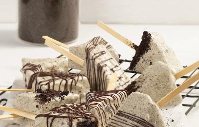 Fotografia em tons de branco em uma bancada de cor branca. No centro, estão os Cakes Pop Cookies’n Cream polvilhados com chocolate. Ao fundo, um recipiente contendo Topping de Negresco. Fotografia em tons de branco em uma bancada de cor branca. No centro, estão os Cakes Pop Cookies’n Cream polvilhados com chocolate. Ao fundo, um recipiente contendo Topping de Negresco.