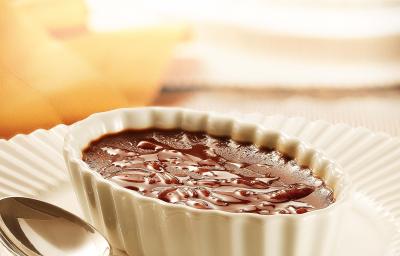 fotografia em tons de branco e marrom de uma bancada vista de frente, contém um prato redondo e branco com um potinho oval branco com um creme de chocolate dentro e uma colher ao lado para servir. fotografia em tons de branco e marrom de uma bancada vista de frente, contém um prato redondo e branco com um potinho oval branco com um creme de chocolate dentro e uma colher ao lado para servir.