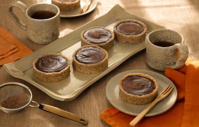 Foto em tons de marrom da receita de torta de ganache de cacau servida em diversas porções sobre uma base de cerâmica retangular e uma sobre um pires de cerâmica, além de duas caneca de mesmo material ao lado Foto em tons de marrom da receita de torta de ganache de cacau servida em diversas porções sobre uma base de cerâmica retangular e uma sobre um pires de cerâmica, além de duas caneca de mesmo material ao lado