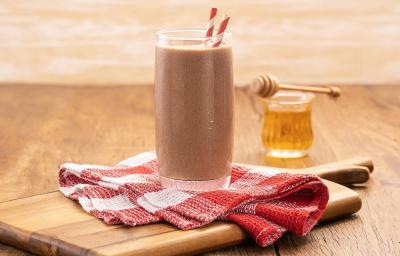 Foto da receita de milkshake proteico com abobrinha e cacau servida em um copo alto e largo com dois canudos listrados, embaixo um pano quadriculado vermelho e branco sobre uma tábua de madeira. Ao fundo um copo do vidro com mel