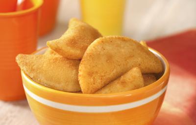Fotografia em tons de amarelo e laranja, ao centro potinho com rissoles fritos, ao lado guardanapo laranja, acima copo amarelo com canudo, no lado esquerdo jarra e copo laranja com um canudo, tudo sobre bancada de madeira em pátina. Fotografia em tons de amarelo e laranja, ao centro potinho com rissoles fritos, ao lado guardanapo laranja, acima copo amarelo com canudo, no lado esquerdo jarra e copo laranja com um canudo, tudo sobre bancada de madeira em pátina.