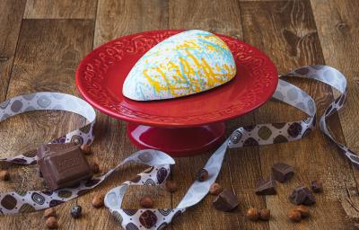 Imagem da receita de uma casca de ovo de páscoa branca, decorada com cores laranja e azul, sobre um prato vermelho, em uma bancada de madeira, decorada com uma fita decorativa, pedaços de chocolate e avelãs Imagem da receita de uma casca de ovo de páscoa branca, decorada com cores laranja e azul, sobre um prato vermelho, em uma bancada de madeira, decorada com uma fita decorativa, pedaços de chocolate e avelãs