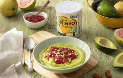 Fotografia em tons de verde de uma bancada verde, sobre ela uma tábua de madeira com um recipiente verde redondo com o creme de abacate e uma colher. Ao lado abacates, um paninho branco, um recipiente com goiabada, goiabas e uma lata de NINHO orgânico. Fotografia em tons de verde de uma bancada verde, sobre ela uma tábua de madeira com um recipiente verde redondo com o creme de abacate e uma colher. Ao lado abacates, um paninho branco, um recipiente com goiabada, goiabas e uma lata de NINHO orgânico.