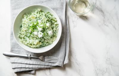 Fotografia em tons de branco e verde de uma bancada branca vista de cima. Contém um pano branco com um recipiente redondo branco e dentro o arroz com o espinafre. Contém um garfo para servir e ao lado esquerdo um copo com vinho.