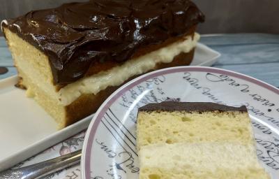 Foto da receita de Boston Pie. Observa-se o bolo retangular com uma camada de creme branco no meio e cobertura de chocolate no plano de fundo. À frente, um prato de sobremesa redondo com uma fatia do bolo. Foto da receita de Boston Pie. Observa-se o bolo retangular com uma camada de creme branco no meio e cobertura de chocolate no plano de fundo. À frente, um prato de sobremesa redondo com uma fatia do bolo.