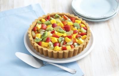 Fotografia em tons de bege, azul, amarelo, vermelho e verde de uma bancada bege vista de cima. Contém um pano azul e por cima um prato redondo branco, com uma torta com frutas picadas (kiwi, manga, morango, uva) por cima. Fotografia em tons de bege, azul, amarelo, vermelho e verde de uma bancada bege vista de cima. Contém um pano azul e por cima um prato redondo branco, com uma torta com frutas picadas (kiwi, manga, morango, uva) por cima.