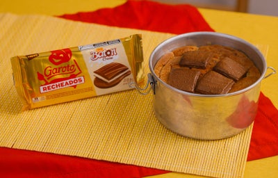 Cookie gigante em lata ao lado da embalagem Garoto Recheados Baton Creme, sobre toalhas vermelha e amarela.