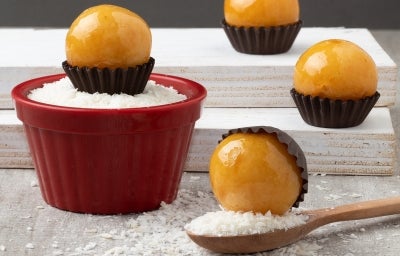 Em cima de uma mesa, pote vermelho com coco ralado, ao lado, balas redondas de caramelo crocante em forminhas marrons. Em cima de uma mesa, pote vermelho com coco ralado, ao lado, balas redondas de caramelo crocante em forminhas marrons.