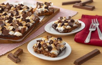 Fotografia em tons de marrom em uma bancada de madeira de cor bege. AO centro, 2 pires contendo os s'mores. Ao fundo, um pano vermelho contendo dois garfos, um pano com bolinhas vermelhas contendo uma tábua mais s'mores e alguns kikat espalhados. Fotografia em tons de marrom em uma bancada de madeira de cor bege. AO centro, 2 pires contendo os s'mores. Ao fundo, um pano vermelho contendo dois garfos, um pano com bolinhas vermelhas contendo uma tábua mais s'mores e alguns kikat espalhados.