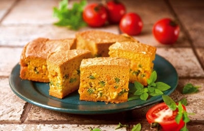 Fotografia de fatias de um bolo salgado em um prato, recheado com vegetais como milho e ervilhas. Tomates frescos e ervas verdes decoram a apresentação.