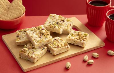 Foto da receita de Fudge de Pistache com TOSTINES Maisena e GALAK. Observa-se uma decoração vermelha com uma tábua de madeira e o fudge cortado por cima. Pistaches decoram a foto.