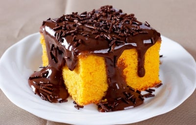 Um pedaço de bolo de cenoura também em formato quadrado, com calda de chocolate escorrendo generosamente pelas laterais e decorado com granulados de chocolate. Ele está servido em um prato branco sobre uma toalha ou superfície marrom.